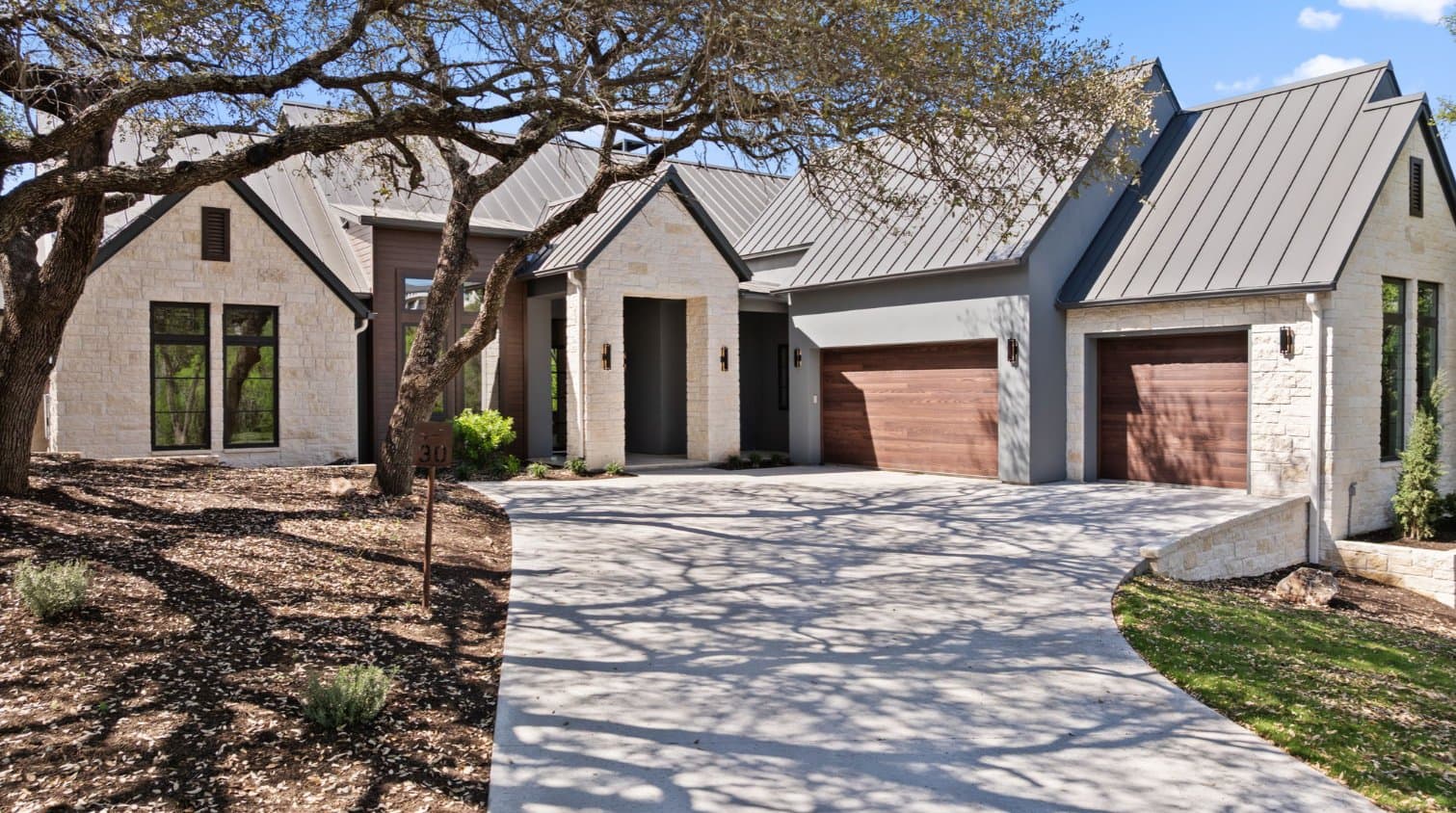 Light stone cottage-style home with steep metal roofs and wood garage doors under an oak