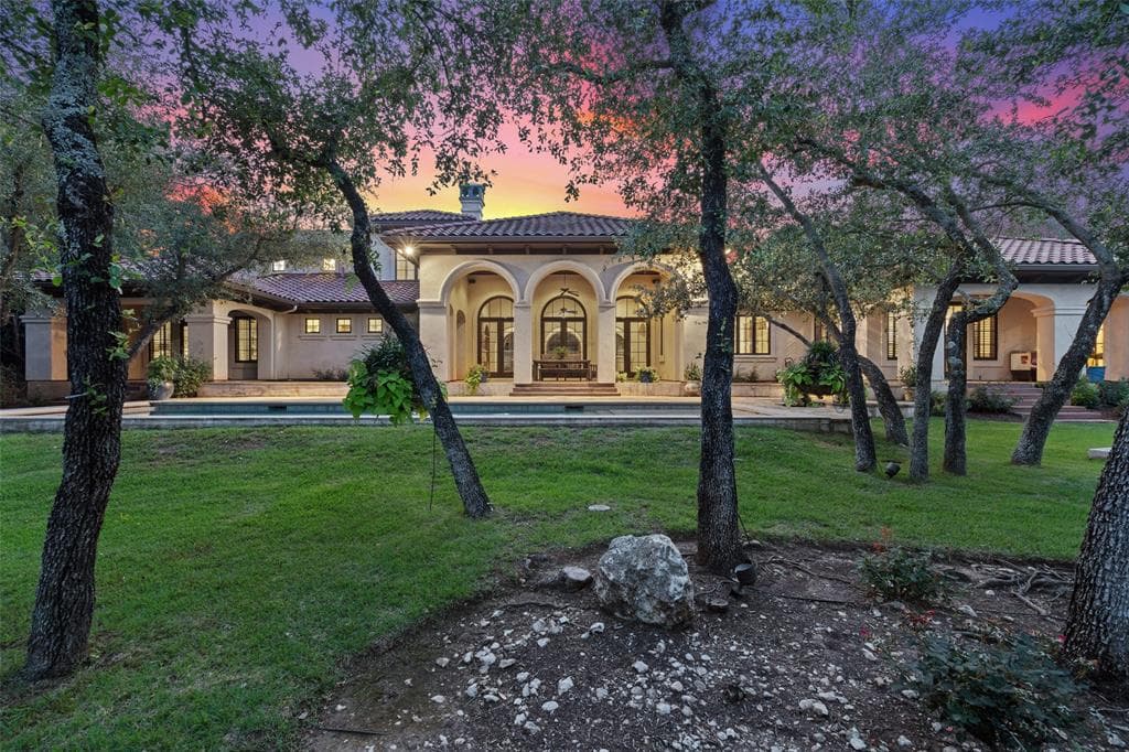 Mediterranean home with arched loggias and a tile roof at dusk, seen through oak trees