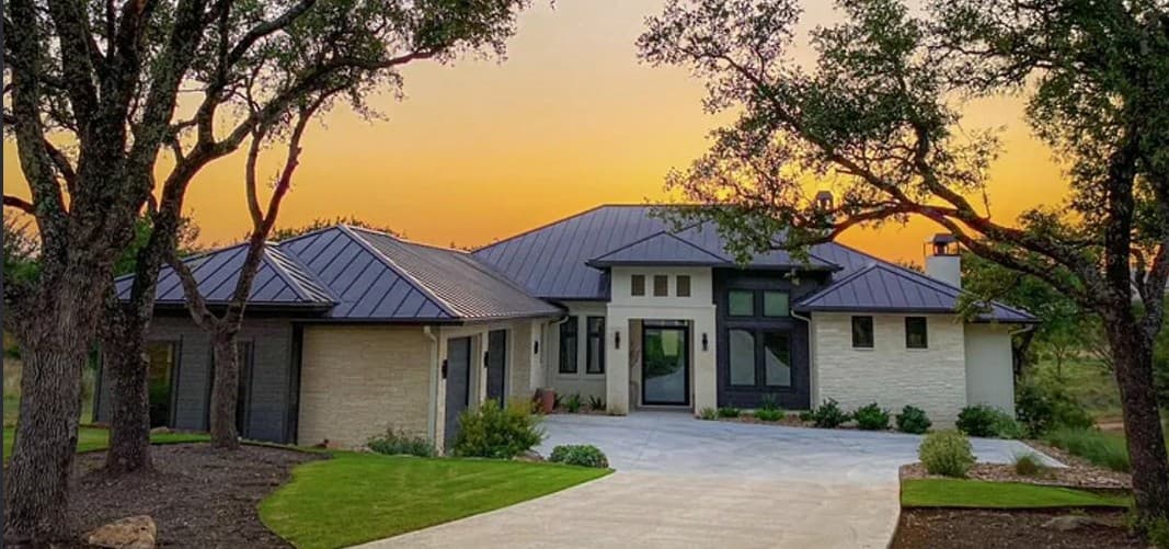 Limestone home with a standing-seam metal roof at sunset, framed by live oaks